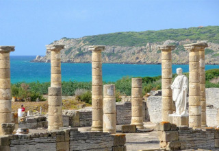 Ruiner af antikke søjler og en statue tæt på havet nær Tarifa, Cádiz, Andalusien, Spanien, med grønne bakker.