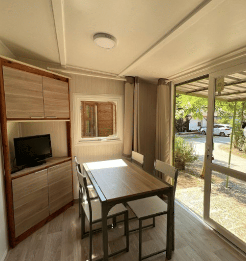 Bright interior at Bungalow Urbasa with dining table, chairs, small TV, and view of greenery outside.