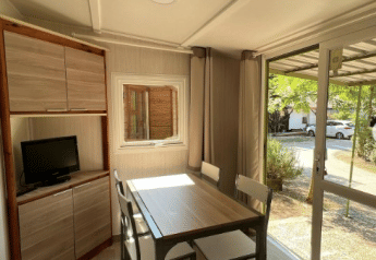Bright interior at Bungalow Urbasa with dining table, chairs, small TV, and view of greenery outside.