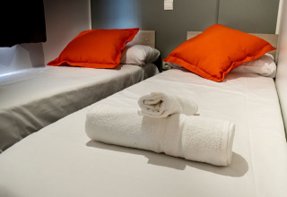 Twee eenpersoonsbedden met witte lakens, oranje kussens en propere handdoeken bij Mobile home Urederra, Camping Etxarri, Spanje.