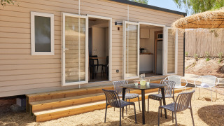 Zona exterior frente a una cabaña moderna con puertas abiertas, mesa, sillas negras y área sombreada de grava.