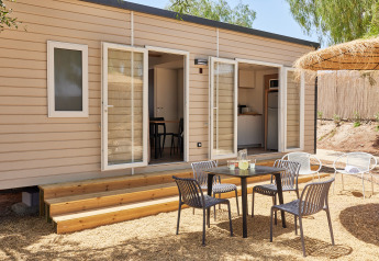 Zona exterior frente a una cabaña moderna con puertas abiertas, mesa, sillas negras y área sombreada de grava.