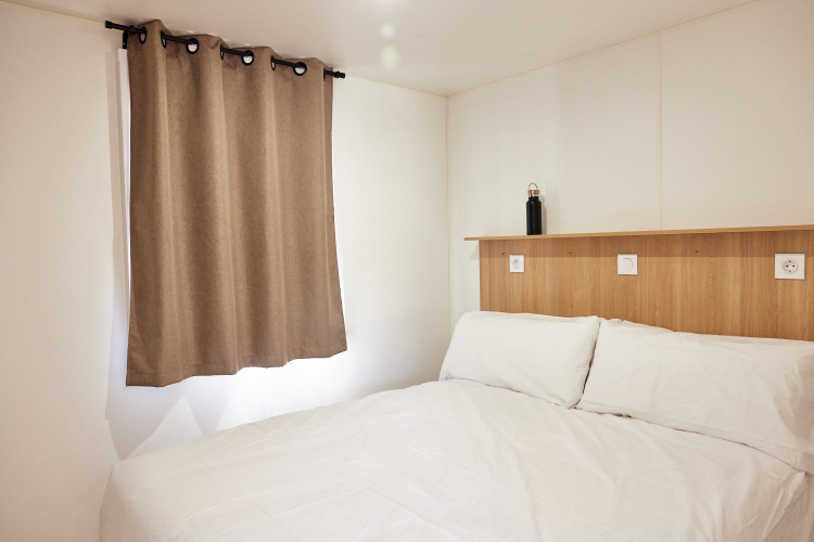 Dormitorio minimalista en cabaña, con cama doble, paredes blancas, cortinas beige y cabecero de madera.