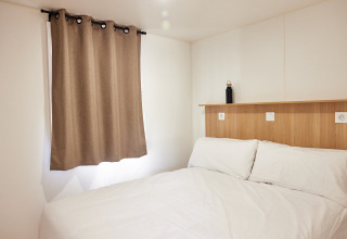 Minimalistisch eingerichtetes Schlafzimmer in einer Hütte mit Doppelbett, weißen Wänden und braunen Vorhängen.