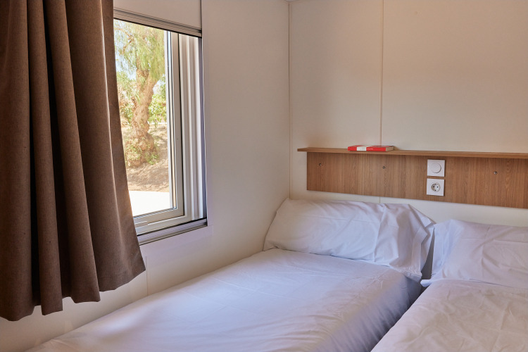Photo of a simple cabin room with two single beds, window with a view, and a wooden shelf above beds.