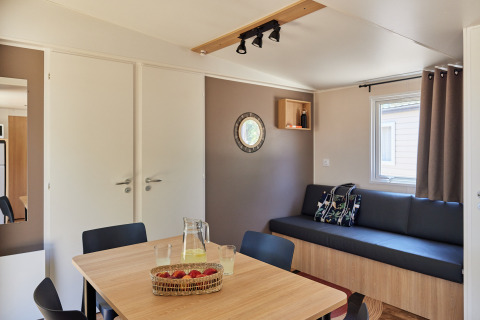 Interior de cabaña moderno y luminoso con mesa de comedor, sofá, ventana, cojines y canasta de frutas.