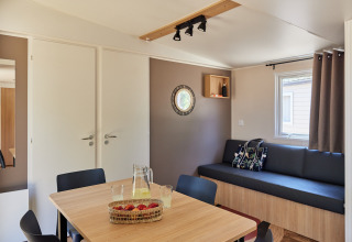 Interior de cabaña moderno y luminoso con mesa de comedor, sofá, ventana, cojines y canasta de frutas.