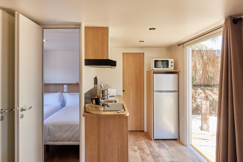 Interior de cabaña moderna y luminosa con cocina, dormitorio, nevera, microondas y ventana al exterior.