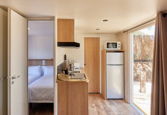 Interior de cabaña moderna y luminosa con cocina, dormitorio, nevera, microondas y ventana al exterior.