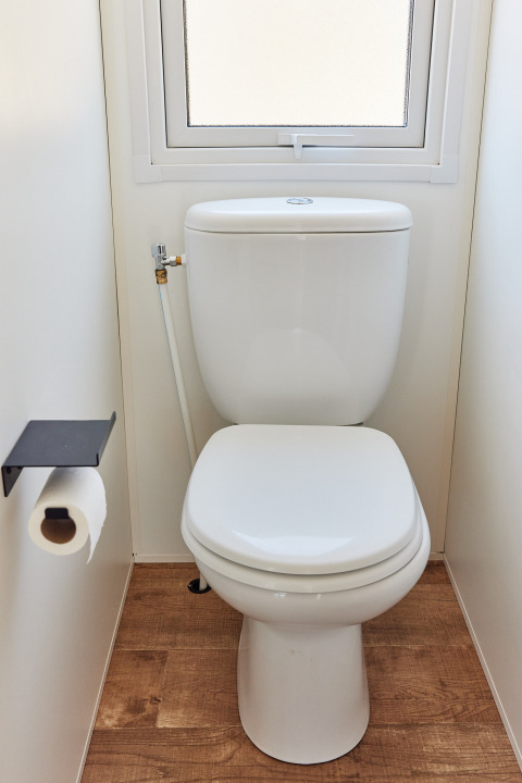 Et rent moderne toilet med toiletpapirholder og vindue i en hytte med trægulv og lyse vægge.