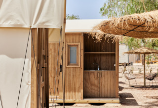 Cocina exterior de madera junto a una tienda safari, bajo sombrillas de paja y árboles altos.