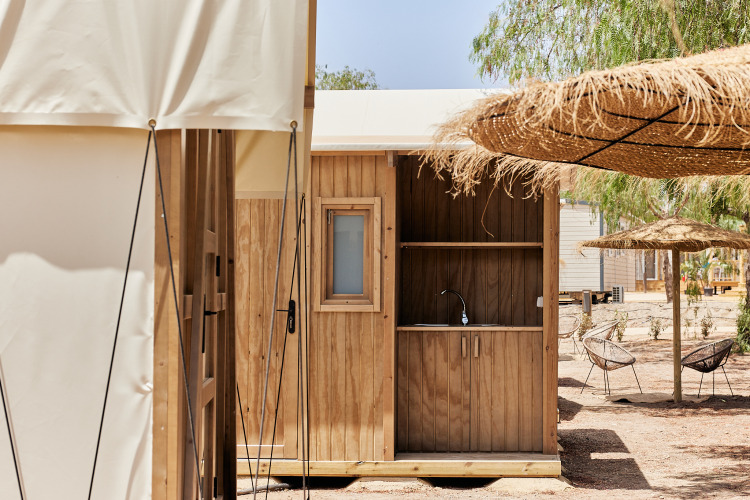 Outdoor wooden kitchen area next to a safari tent, shaded by large straw umbrellas and tall trees.