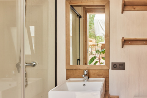 Baño en tienda safari con lavabo blanco, espejo con marco de madera, ducha y vista al exterior.