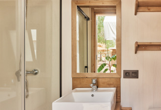 Baño en tienda safari con lavabo blanco, espejo con marco de madera, ducha y vista al exterior.