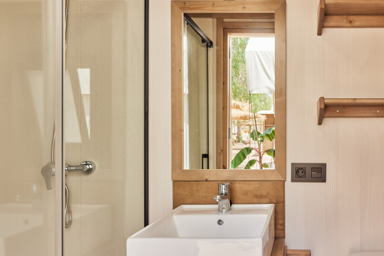 Safari tent bathroom with a white sink, wooden-framed mirror, shower, and view of greenery outside.
