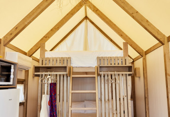 Interno di una tenda safari con letti a castello in legno, scala, frigorifero e lampada intrecciata.