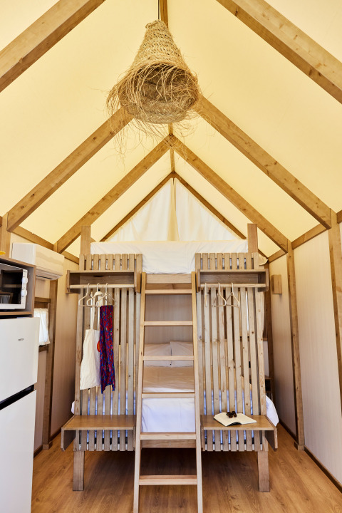Interno di una tenda safari con letti a castello in legno, scala, frigorifero e lampada intrecciata.