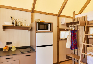Keukentje in een safaritent met koelkast, microgolfoven, planken, keukengerei en een stapelbed aanwezig.