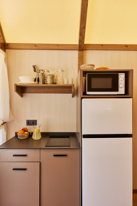Compact kitchenette in a safari tent with stovetop, fridge, microwave, shelves, and fruit basket.