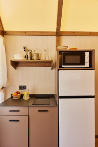 Compact kitchenette in a safari tent with stovetop, fridge, microwave, shelves, and fruit basket.