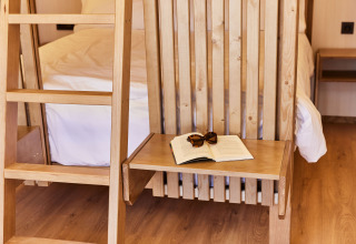 Interior de tienda safari con litera de madera, mesa, libro abierto y gafas de sol, ambiente acogedor.