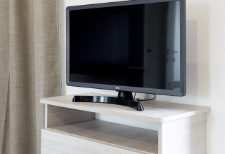 Televisor de pantalla plana sobre mueble de madera clara junto a cortina beige en Bungalow Confort Baconé, TAIGA Delta de l'Ebre.