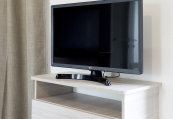 Televisor de pantalla plana sobre mueble de madera clara junto a cortina beige en Bungalow Confort Baconé, TAIGA Delta de l'Ebre.