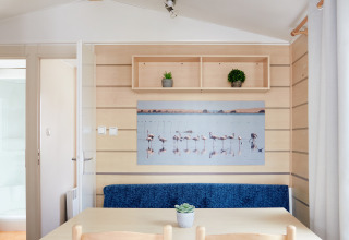 Interior de Bungalow Confort Delta con mesa de comedor, dos sillas, banco azul y cuadro de flamencos en la pared.