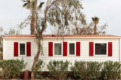 Bungaló blanco con contraventanas rojas en TAIGA Delta de l'Ebre, España, rodeado de árboles y arbustos.