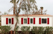 Bungaló blanco con contraventanas rojas en TAIGA Delta de l'Ebre, España, rodeado de árboles y arbustos.
