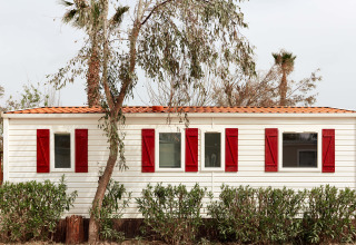 Bungaló blanco con contraventanas rojas en TAIGA Delta de l'Ebre, España, rodeado de árboles y arbustos.