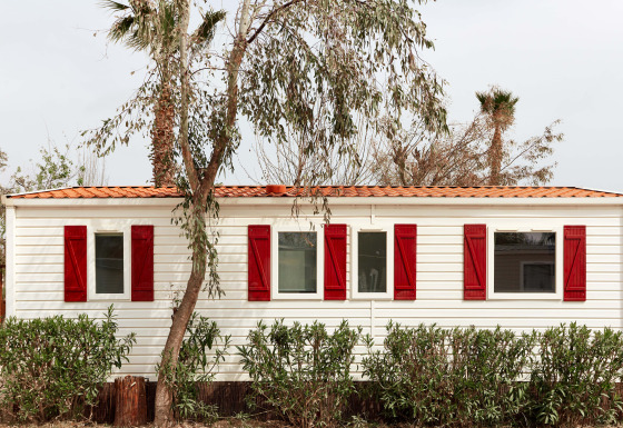 Bungaló blanco con contraventanas rojas en TAIGA Delta de l'Ebre, España, rodeado de árboles y arbustos.