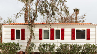 Bungaló blanco con contraventanas rojas en TAIGA Delta de l'Ebre, España, rodeado de árboles y arbustos.