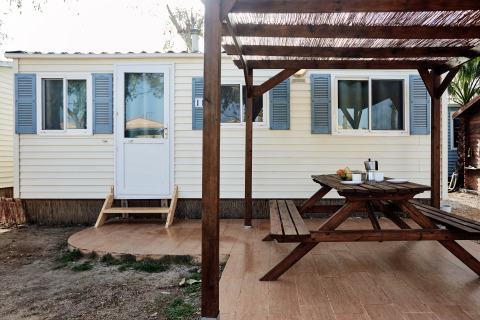 Terraza exterior con mesa y bancos de madera frente a una cabaña con contraventanas azules en Bungalow Essential Flamenc.