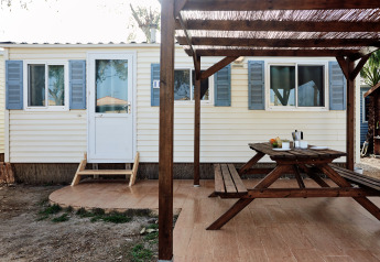 Terraza exterior con mesa y bancos de madera frente a una cabaña con contraventanas azules en Bungalow Essential Flamenc.