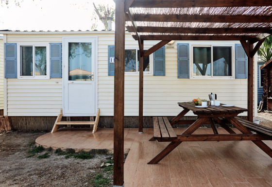 Terraza exterior con mesa y bancos de madera frente a una cabaña con contraventanas azules en Bungalow Essential Flamenc.