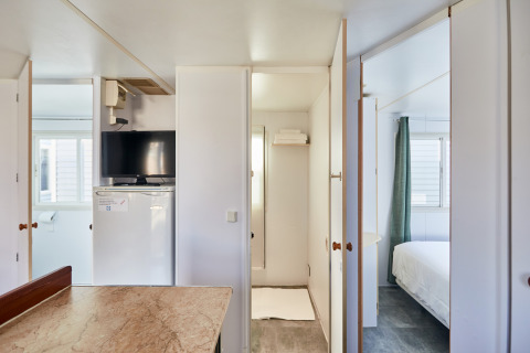 Innenansicht eines hellen Bungalows mit Fernseher, Kühlschrank, Tisch sowie Zugang zu Schlafzimmer und Bad.