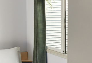 Habitación minimalista con cama blanca, cortina verde y ventana en Bungalow Essential Flamenc, TAIGA Delta de l'Ebre.