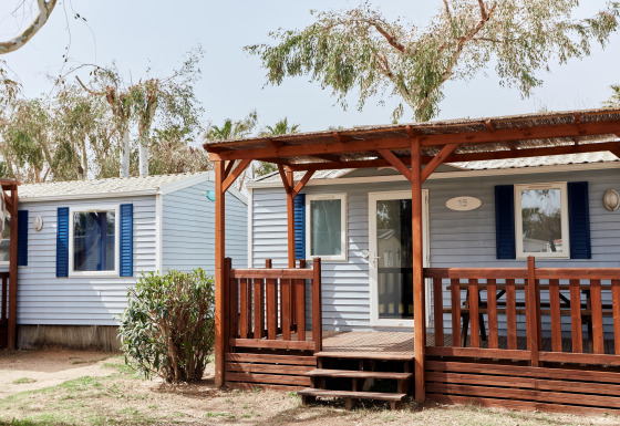 Buitenaanzicht van Bungalow Confort Daurada met houten terras, blauwe luiken en bomen op de achtergrond.