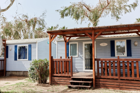 Buitenaanzicht van Bungalow Confort Daurada met houten veranda, blauwe luiken en bomen op de achtergrond.