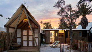Glamping Cabin-Tent Senyoret bei Sonnenuntergang mit stilvollen Holzhütten, Palmen und Liegestühlen.