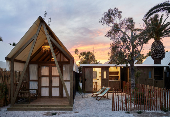 Glamping Cabin-Tent Senyoret bij zonsondergang met houten cabines, palmbomen en strandstoelen buiten.