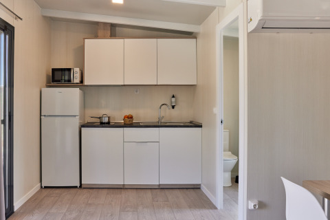 Pequeña cocina glamping con armarios blancos, nevera, microondas y acceso visible al baño.