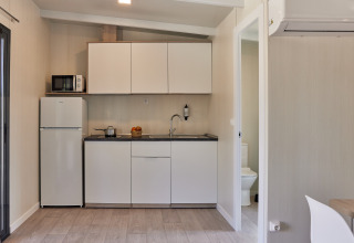 Pequeña cocina glamping con armarios blancos, nevera, microondas y acceso visible al baño.
