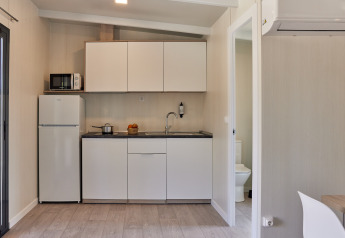 Pequeña cocina glamping con armarios blancos, nevera, microondas y acceso visible al baño.