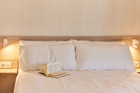 Cama acogedora con almohadas blancas, luces de lectura y libro abierto en alojamiento glamping moderno.