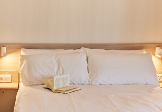 Cama acogedora con almohadas blancas, luces de lectura y libro abierto en alojamiento glamping moderno.