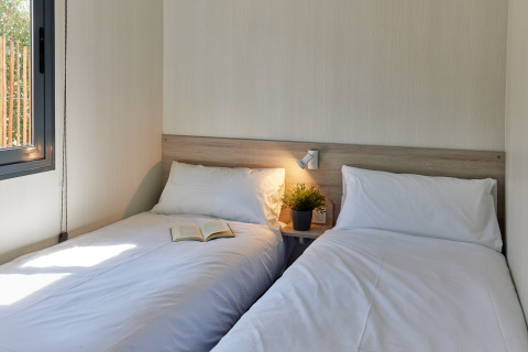 Deux lits simples avec du linge blanc dans une chambre de glamping lumineuse, livre et plante sur la table de nuit.