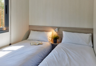 Twee enkele bedden met wit linnengoed in een lichte glampingkamer, boek en plantje op het nachtkastje.