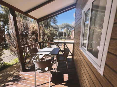 Porche de madera con mesa y sillas en el Bungalow Confort Poniente, TAIGA Conil, España, rodeado de árboles.